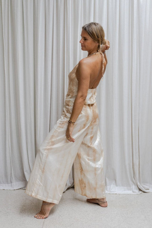 A woman wearing a beige tea-dye elegant silk romper with a halter-style neckline and wide-leg silhouette, standing with a white backdrop in the background, side view.
