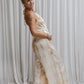 A woman wearing a beige tea-dye elegant silk romper with a halter-style neckline and wide-leg silhouette, standing with a white backdrop in the background. Side view 2