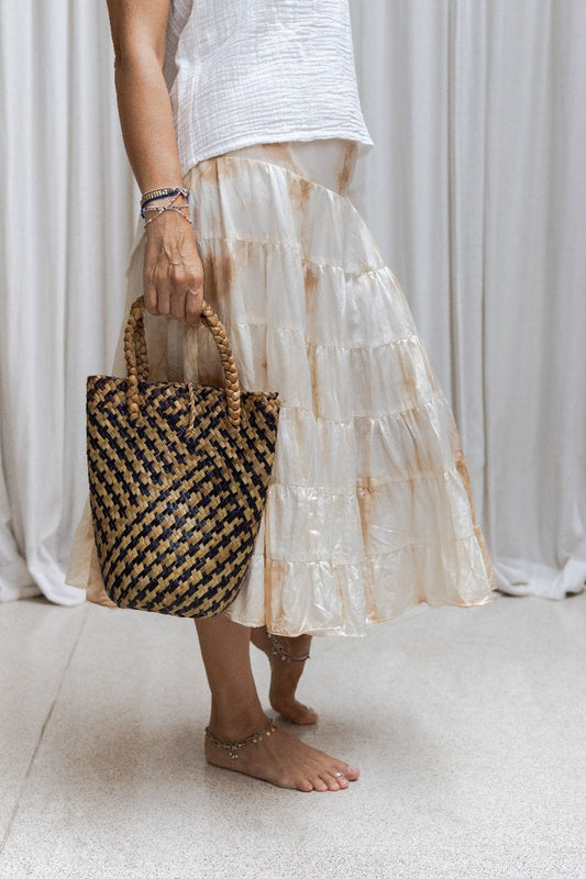 A woman wearing a tea dye silk skirt with a gathered tiered design and an elasticized waistband, paired with a white sleeveless top. Holding a small bag 