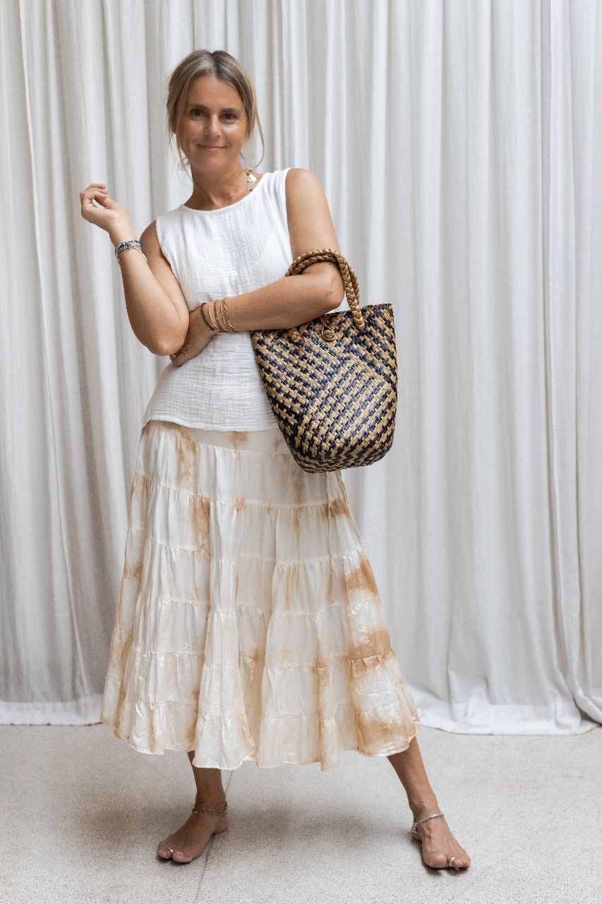 A woman wearing a white sleeveless top and the PRIYA Tea Dye Silk Skirt with an elasticized waistband. She is holding a patterned handbag and standing against a white backdrop.
