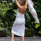 A woman wearing a white linen dress with a halter neckline and open-back silhouette, standing on a wooden deck with greenery in the background. Back view.
