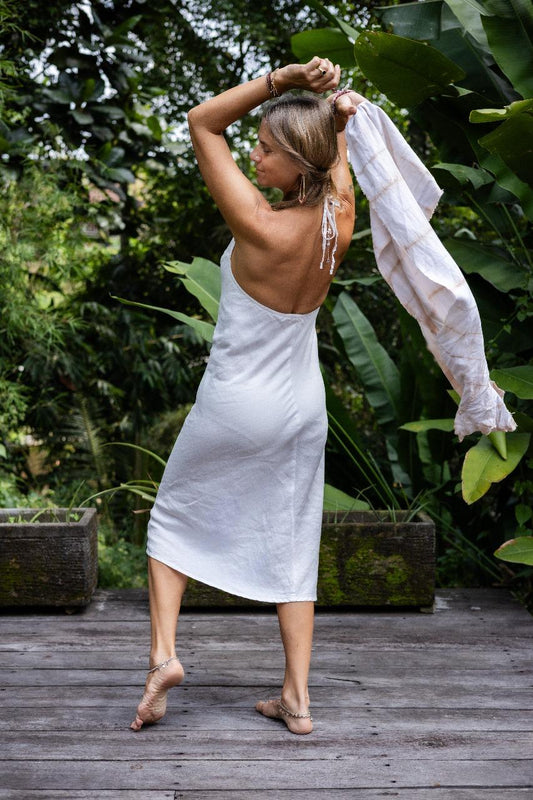 A woman wearing a white linen dress with a halter neckline and open-back silhouette, standing on a wooden deck with greenery in the background. Back view.