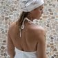 A woman wearing a white linen dress,  with a halter neckline and open-back silhouette, standing in front of a textured wall. Detail.