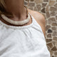 A woman wearing a white linen dress,  with a halter neckline and open-back silhouette, standing in front of a textured wall. Front detail.