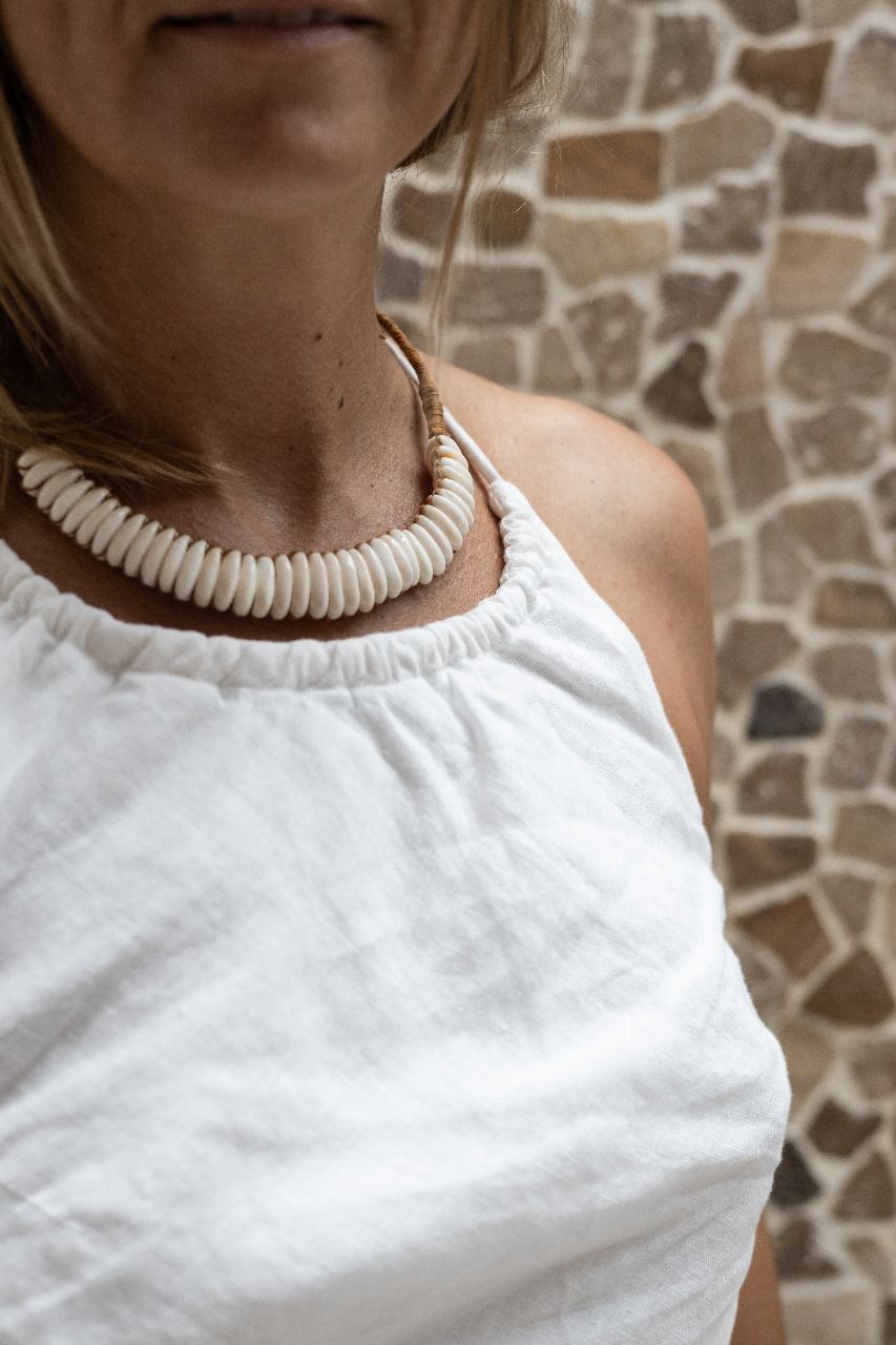 A woman wearing a white linen dress,  with a halter neckline and open-back silhouette, standing in front of a textured wall. Front detail.