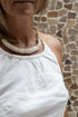 A woman wearing a white linen dress,  with a halter neckline and open-back silhouette, standing in front of a textured wall. Front detail.