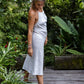 A woman wearing a white linen dress,  with a halter neckline and open-back silhouette, paired with linen pants and standing on a wooden deck in front of a greenery.