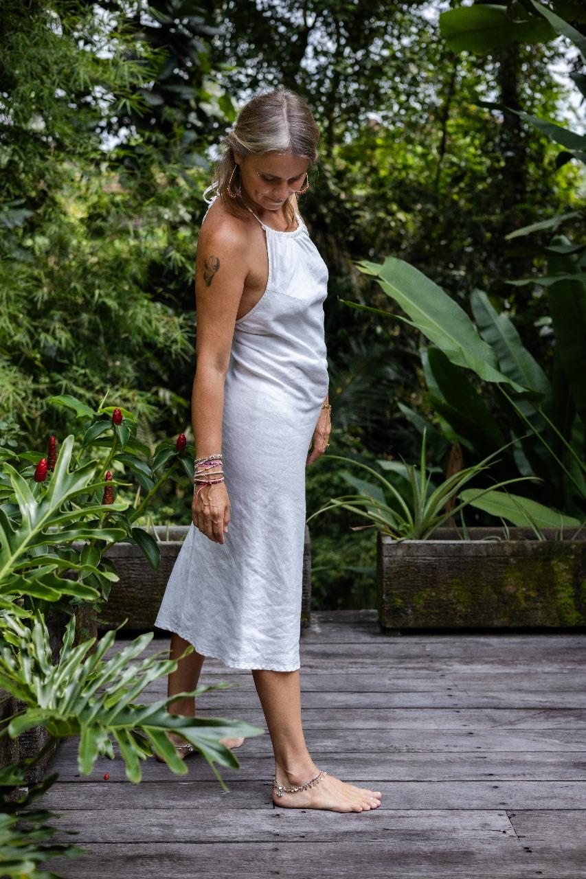 A woman wearing a white linen dress,  with a halter neckline and open-back silhouette, paired with linen pants and standing on a wooden deck in front of a greenery.