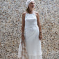 A woman wearing a white linen dress,  with a halter neckline and open-back silhouette, paired with white linen pants and standing in front of a textured wall.