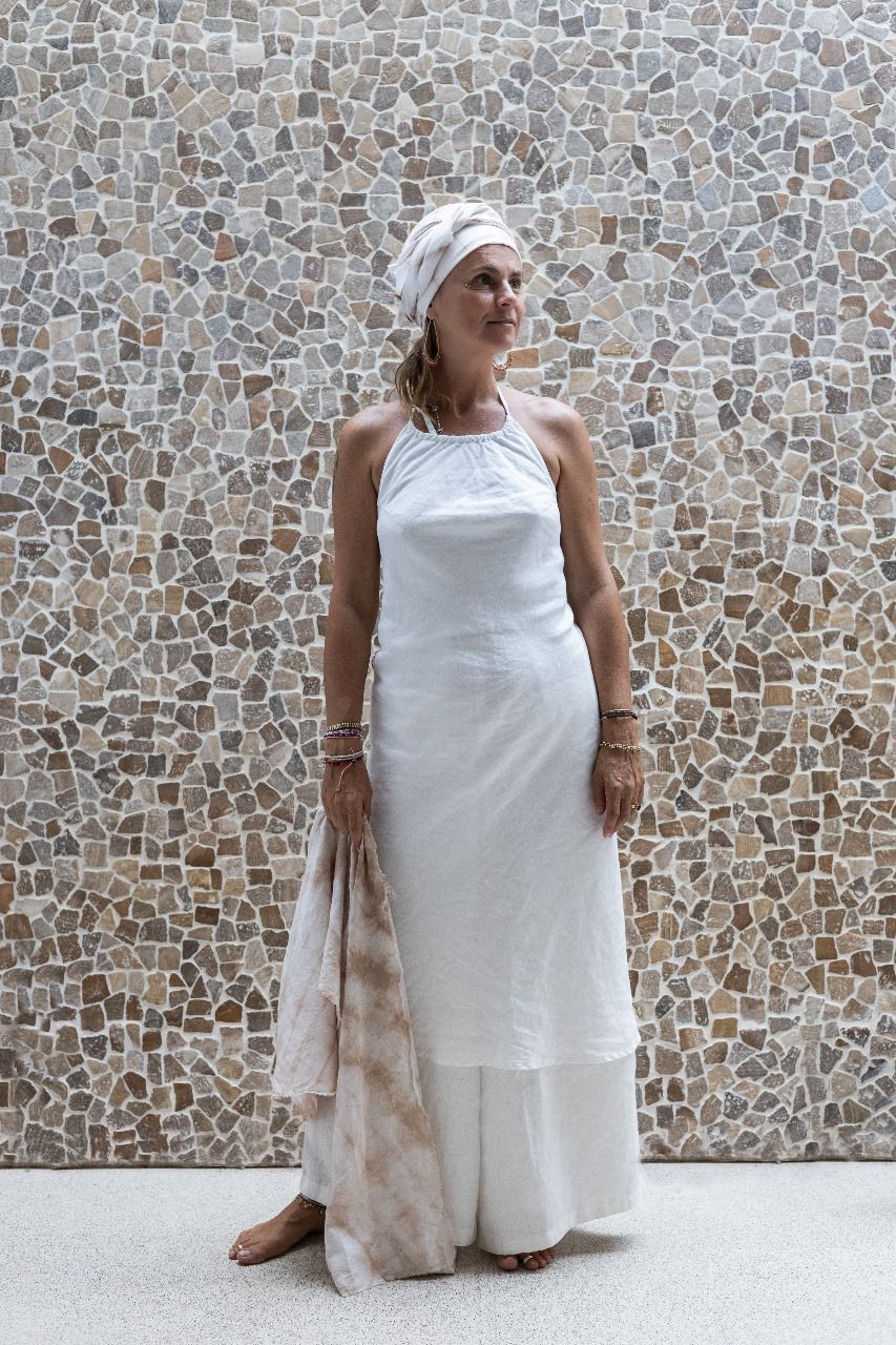 A woman wearing a white linen dress,  with a halter neckline and open-back silhouette, paired with white linen pants and standing in front of a textured wall.