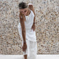 A woman wearing a white linen dress,  with a halter neckline and open-back silhouette, paired with linen pants and standing in front of a textured wall.