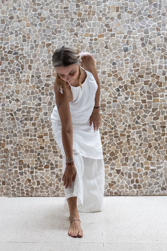 A woman wearing a white linen dress,  with a halter neckline and open-back silhouette, paired with linen pants and standing in front of a textured wall.