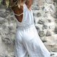 A person wearing a white sleeveless romper with wide legs and a V-neckline, standing with a stone wall in the background. Back view with a deep V 