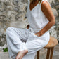 A person wearing a white sleeveless romper with wide legs and a V-neckline, seated on a wooden stool with a stone wall in the background.