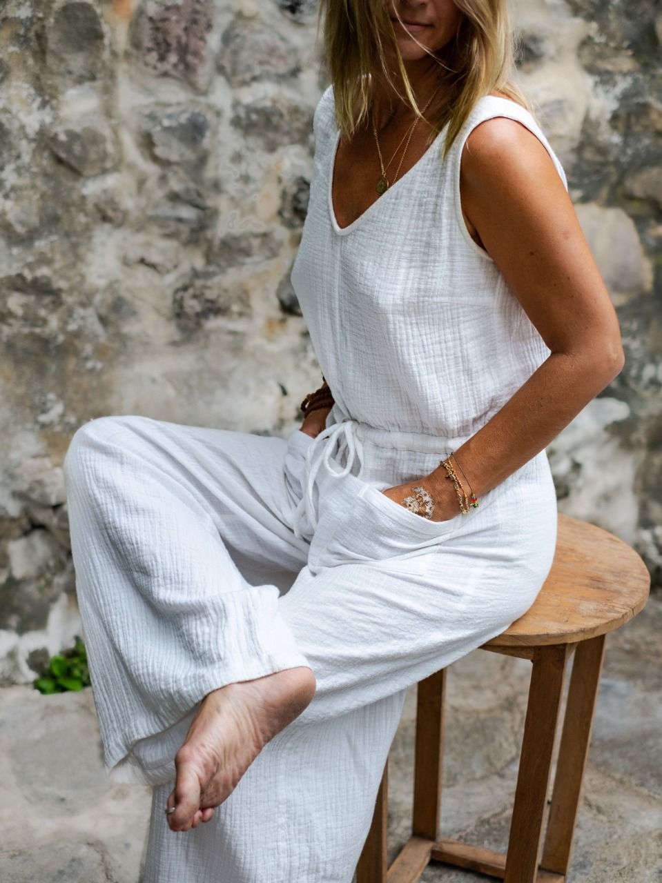 A person wearing a white sleeveless romper with wide legs and a V-neckline, seated on a wooden stool with a stone wall in the background.