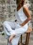 A person wearing a white sleeveless romper with wide legs and a V-neckline, seated on a wooden stool with a stone wall in the background.