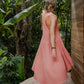 A woman from back, in a rose color  asymmetric cotton dress with a sleeveless design standing near a wooden structure and lush greenery in the background.