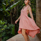 A woman in a rose color asymmetric cotton dress with a sleeveless design standing near a wooden structure and lush greenery in the background.