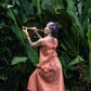 A woman wearing a sleeveless light terracotta top and a matching skirt standing in front of a lush green jungle background and playing a flute. Back view.