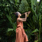 A woman wearing a sleeveless light terracotta top and a matching skirt standing in front of a lush green jungle background and playing a flute. Side view.
