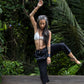 A woman standing against a lush green backdrop, wearing black linen hipster pants and a white cotton bra. All Shivani Divine Wear.