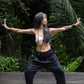 A woman standing against a lush green backdrop, wearing black linen hipster pants and a white cotton bra, front view. All Shivani Divine Wear.