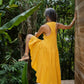 A woman from the back in a yellow asymmetric cotton dress with a sleeveless design standing with her one leg raised, near a wooden structure and lush greenery in the background.