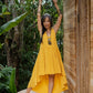 A woman in a yellow asymmetric cotton dress with a sleeveless design standing with her arms raised in the air, near a wooden structure and lush greenery in the background.