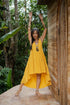 A woman in a yellow asymmetric cotton dress with a sleeveless design standing with her arms raised in the air, near a wooden structure and lush greenery in the background.