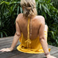 A woman wearing a white, open back tie cotton dress with a texture, sitting on a wooden deck, back view