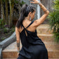 A woman standing on outdoor steps, in a black flowing linen dress, with arms extended gracefully.