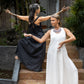 Two women standing on outdoor steps, one in a black and the other in a white flowing linen dress, both with arms extended gracefully.