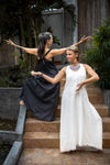 Two women standing on outdoor steps, one in a black and the other in a white flowing linen dress, both with arms extended gracefully.