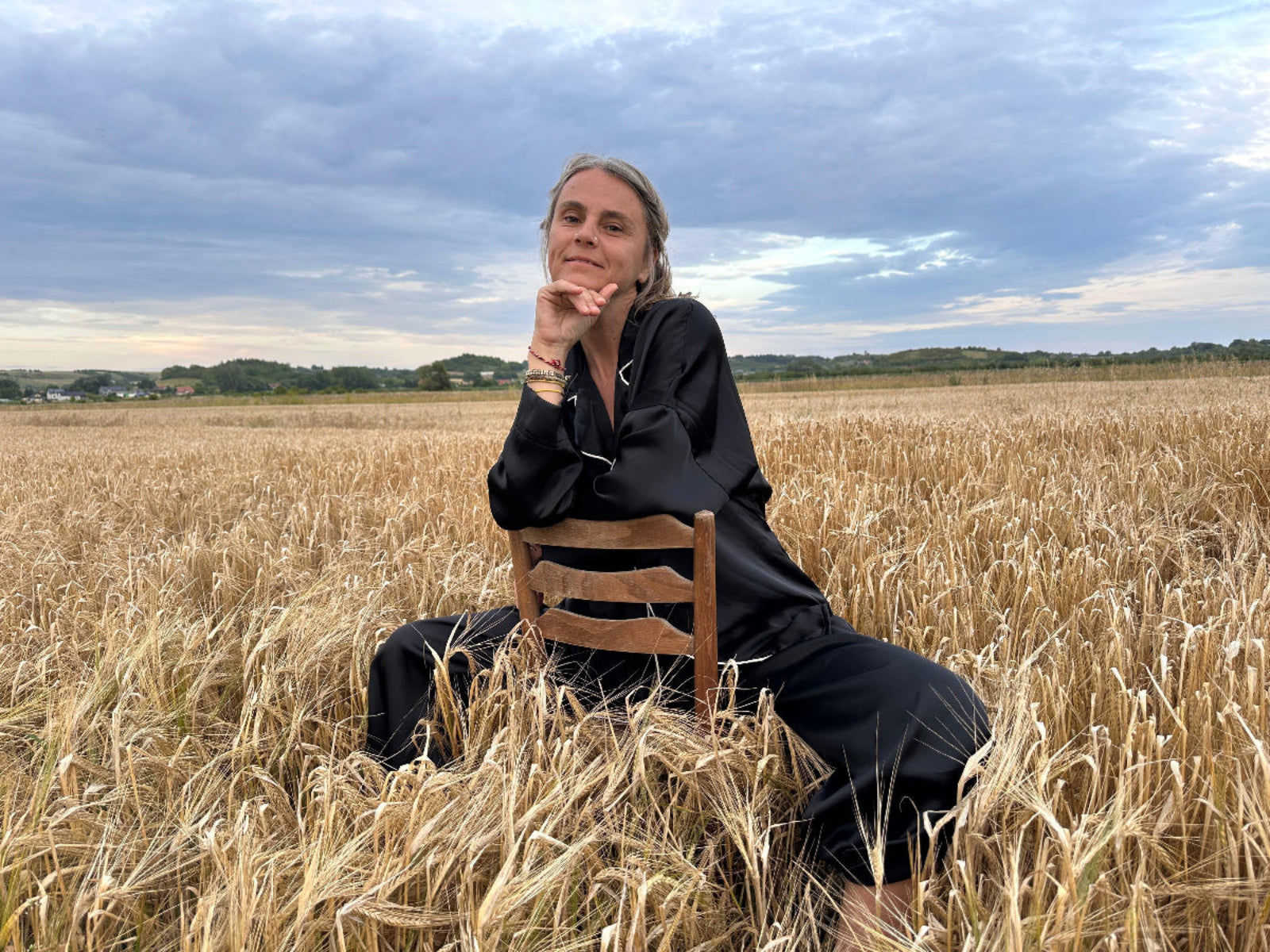 A woman wearing shivani Silk pajama set and sitting on a chair in a wheat field with a cloudy sky.