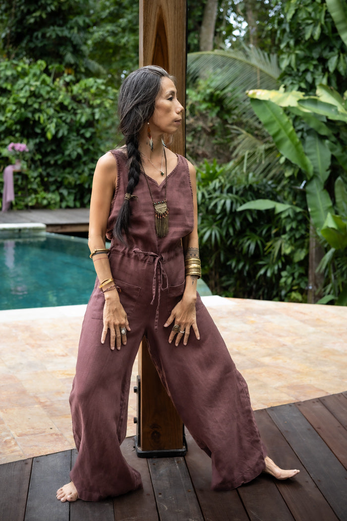 A woman wearing a brown linen romper with wide legs and a V-neckline, standing  in a lush green pool side landscape.