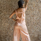 A woman, her back turned to the camera. wearing a rose pink linen romper with wide legs and a deep V-back, standing against a textured background.