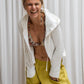 A woman wearing a white hoodie with a zipper and a flowing hood, paired with yellow pants. and viscose flowery bra