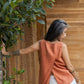 A person wearing a sleeveless crew-neck top in a warm rose color, with an open-back design, standing with their back towards the camera against a wooden  wall/door.