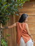 A person wearing a sleeveless crew-neck top in a warm rose color, with an open-back design, standing with their back towards the camera against a wooden  wall/door.