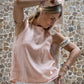 A person wearing a sleeveless crew-neck top in a warm rose color, standing against a textured wall.