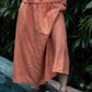 A person wearing a rose-hued skirt with a flowing silhouette, standing near a pool with greenery in the background.