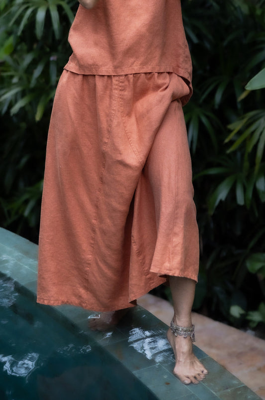 A person wearing a rose-hued skirt with a flowing silhouette, standing near a pool with greenery in the background.