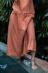 A person wearing a rose-hued skirt with a flowing silhouette, standing near a pool with greenery in the background.