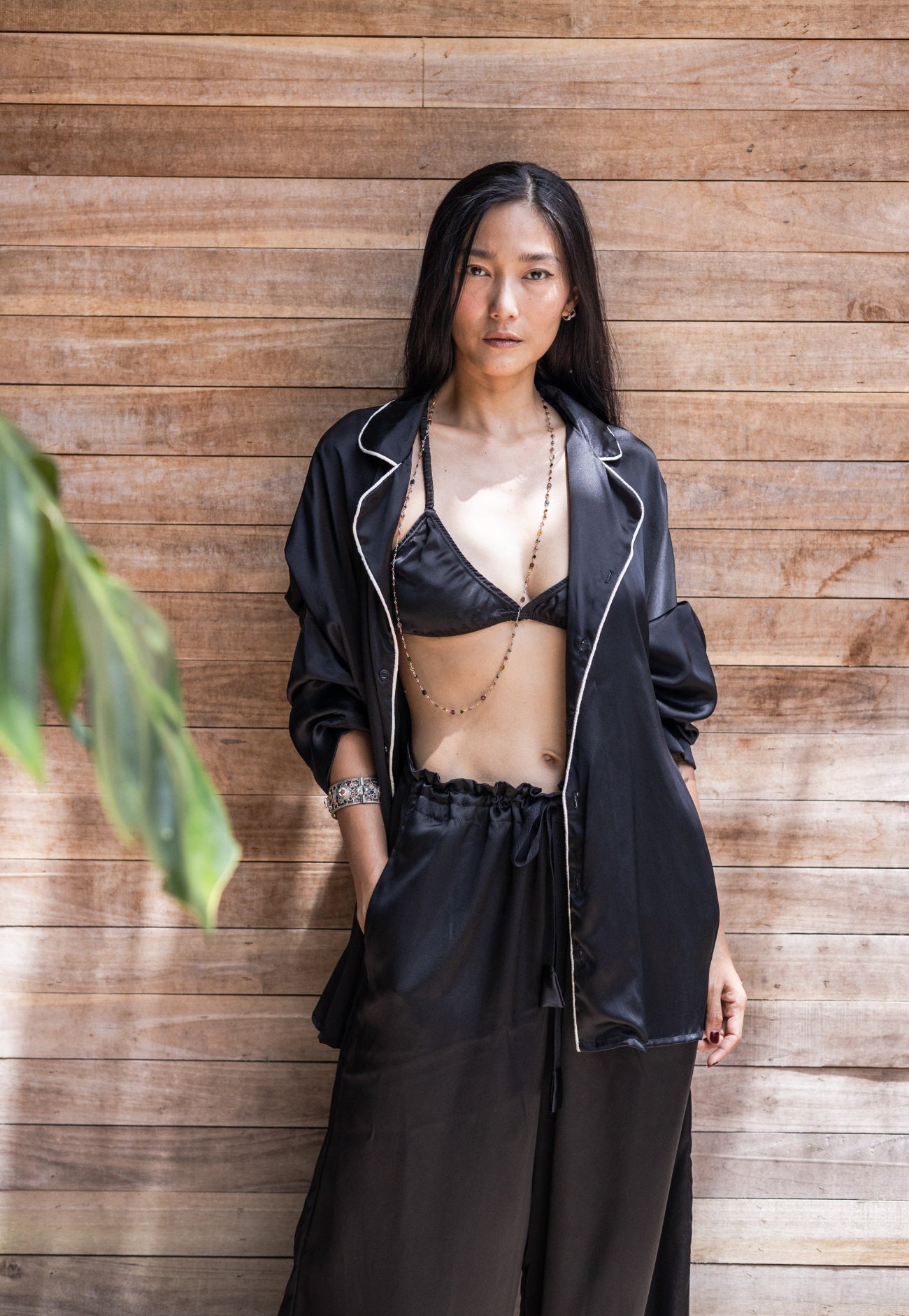Woman wearing a black ssilk pajamas standing against a wooden wall. Top unbuttoned showing silk bra