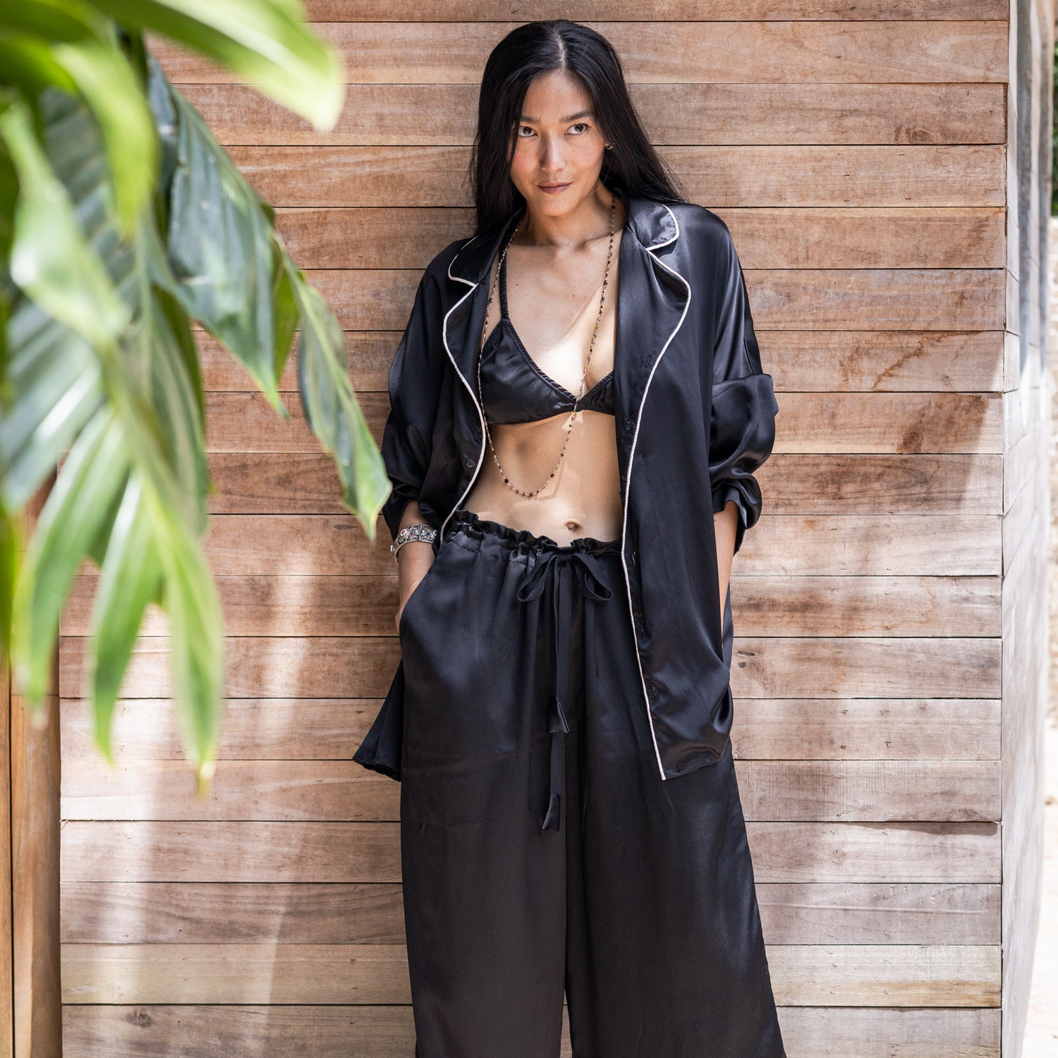 Woman wearing a black silk pajama standing against a wooden wall.