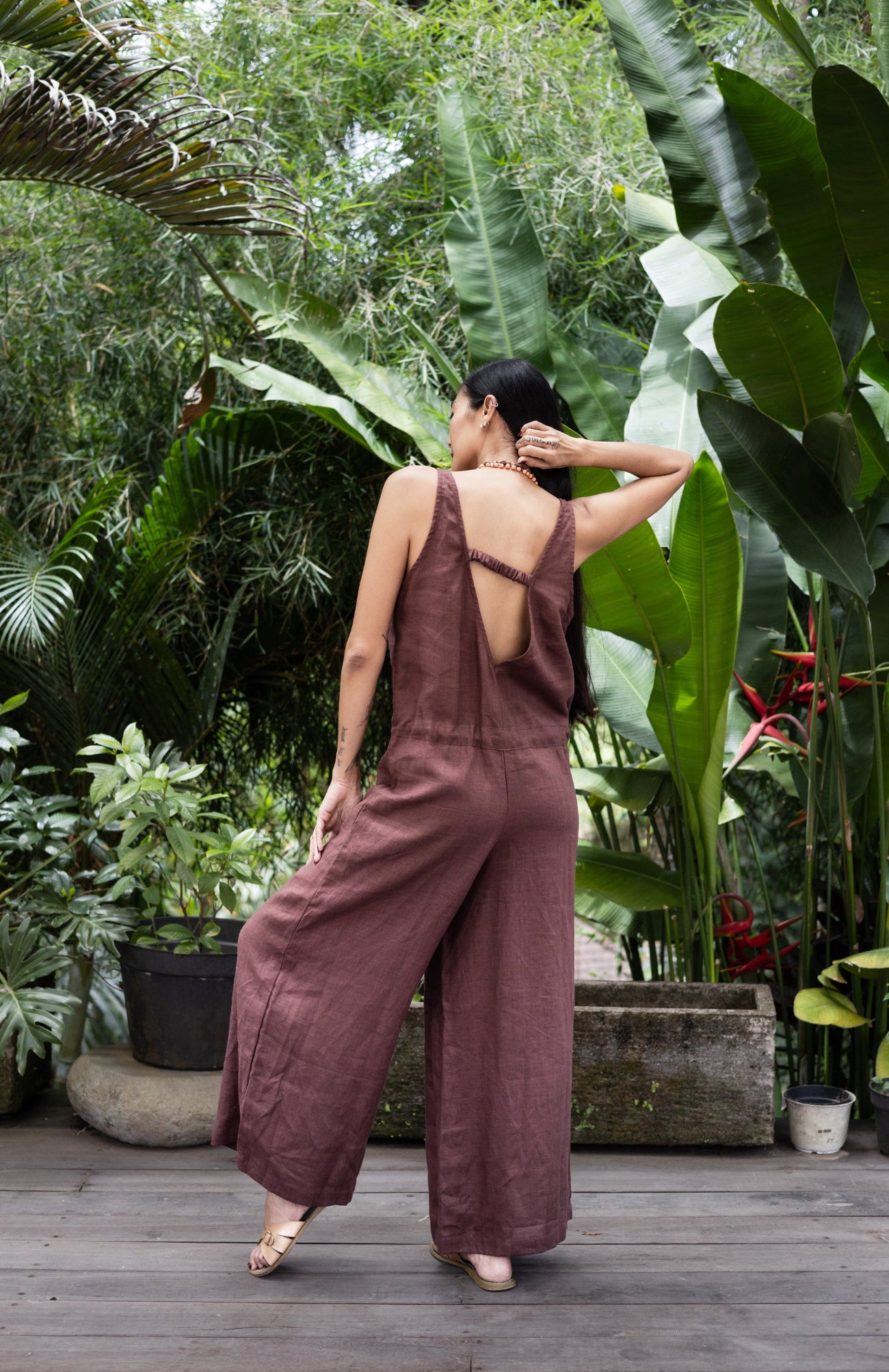 Woman in a brown linen jumpsuit standing in a lush, green garden.
