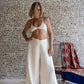 A woman standing on a wooden deck wearing white linen pants and a matching color cotton bra, with stone wall in the background. All Shivani Divine Wear.
