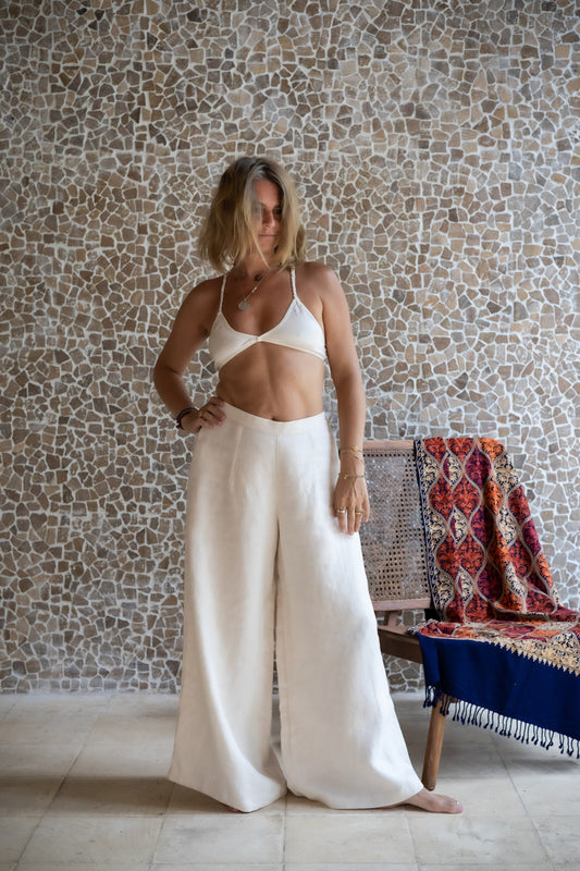 A woman standing on a wooden deck wearing white linen pants and a matching color cotton bra, with stone wall in the background. All Shivani Divine Wear.