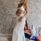 A woman standing on a wooden deck wearing white linen pants and a matching color cotton bra, with stone wall and chair in the background. All Shivani Divine Wear.